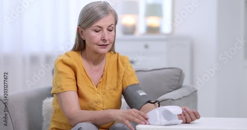Senior woman measuring blood pressure on armchair at white table indoors. Camera moving