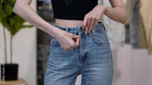 Young Caucasian white woman fastens fitted blue jeans, turns and strokes hips in home wardrobe interior. Concept of flattering denim fit with slim waist, feminine confidence and modern casual clothing