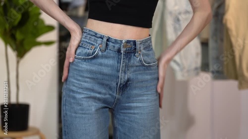 Slender woman zips and buttons high waist blue jeans, smoothing hips and turning in home interior. Concept of flattering denim fit, slim figure, body confidence, style and fashion fitting