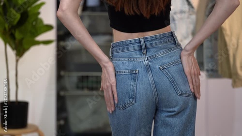 Slender woman in high waist blue jeans strokes hips and turns in home interior, showing flattering fit. Concept of body confidence, slim silhouette, denim styling, flat stomach and everyday fashion