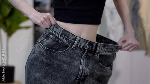 Slim woman joyfully sways hips while stretching big jeans sideways after weight loss, home interior. Concept of healthy lifestyle, diet success, wellness progress, body transformation and motivation