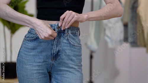 Young slim Caucasian woman buttoning high-waisted jeans, turning and smoothing hips in home dressing area, showing flat stomach. Concept of denim fit check, body confidence and casual fashion styling