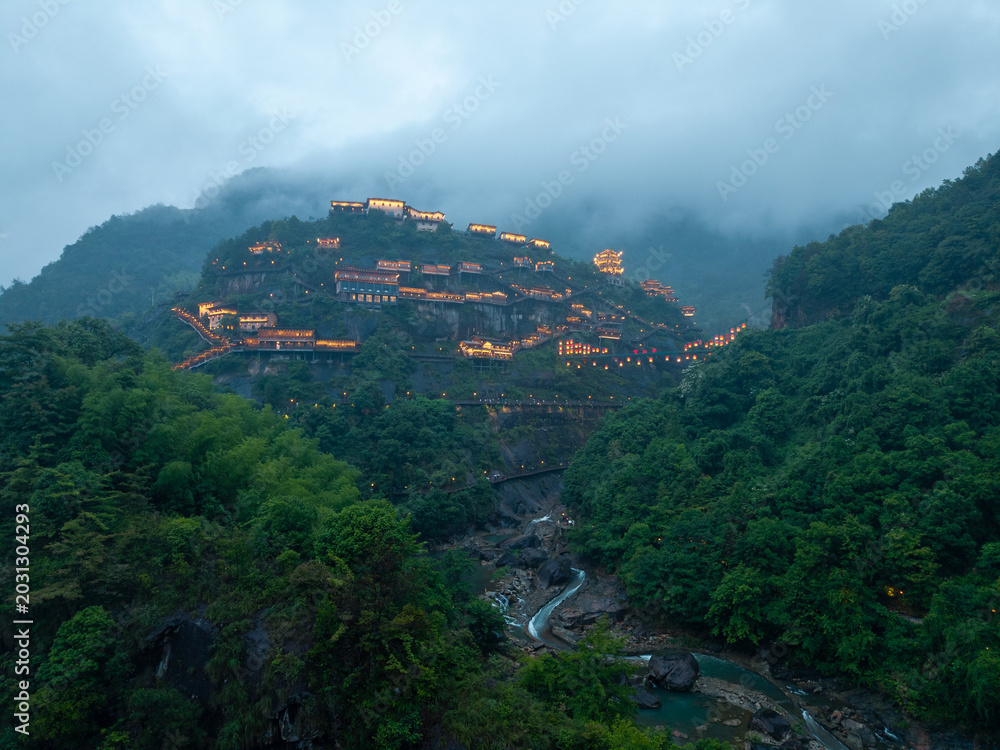 Fototapeta premium A view of the illuminated ancient buildings on the cliffs of Wangxian Valley in Jiangxi China during a misty evening