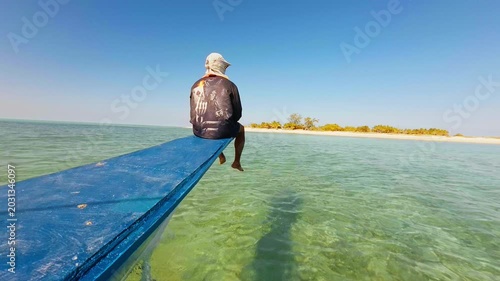 A local boatman or tourist sitting on the front plank of a traditional vessel sailing across clear turquoise ocean water.