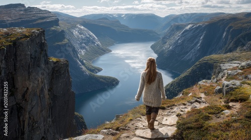 Rear view of a young woman in warm sweater standing on a rocky cliff, looking over a deep blue fjord surrounded by steep mountains on a clear day