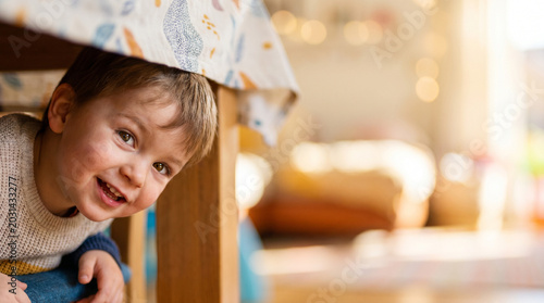 Joyful child hiding under a kitchen table with colorful cloth, smiling in warm sunlight, cozy home interior background