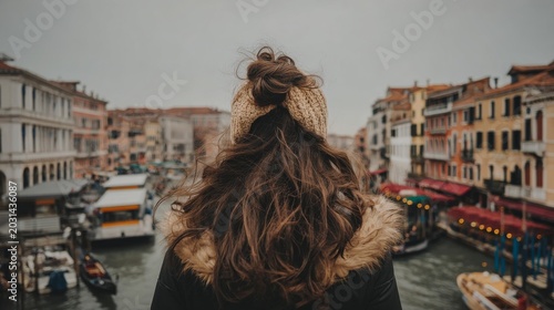 Rear view of a young woman in warm coat and knit hat standing on a bridge, looking at a canal with boats and colorful buildings on a cloudy day in Venice, Italy