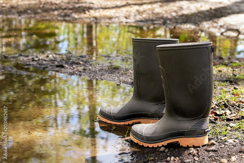 Foto Pair of black rubber boots standing near the muddy puddle in the bushy backyard in spring wet weather