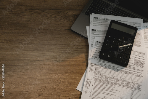 Calculator and pen placed on tax forms beside laptop on wooden desk for checking tax documents, accounting review, financial calculation, expense reporting, and tax filing workflow.