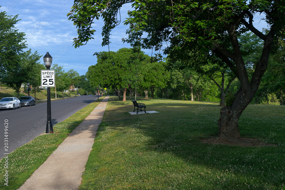 Naklejka premium A speed limit 25 sign and empty metal park bench sit along Carroll Parkway in Baker Park, Frederick, Maryland. Mature trees frame the roadside scene under an overcast morning sky.
