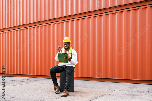 Logistics worker in a hard hat sits by a shipping container, using a radio and checking a clipboard at an industrial yard.