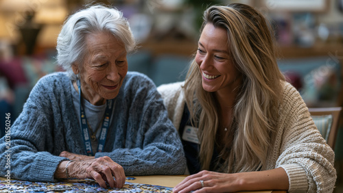 Elderly woman with early-stage dementia sits at a table working on a puzzle with a caregiver in a cozy room during afternoon hours