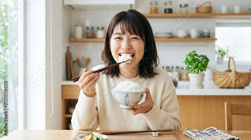 Happy Japanese woman eating a large mouthful of white rice from a bowl in a bright modern kitchen