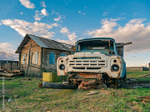 Foto Old blue truck sits in field with wood logs under its front near abandoned wooden structure