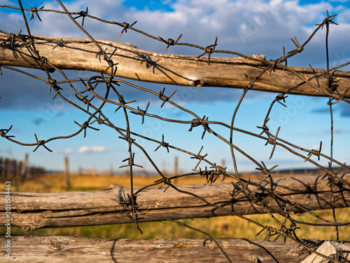 Bild auf Leinwand Barbed wire on wooden fence against blue sky and open field in rural area during