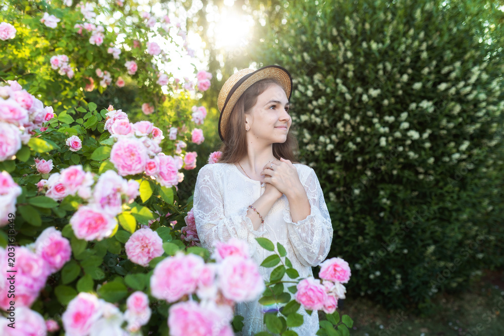 Fototapeta premium Beautiful girl in white dress and straw hat near blooming pink roses in a summer garden. Cute child with long hair with flowers outdoors. Romantic portrait with floral background