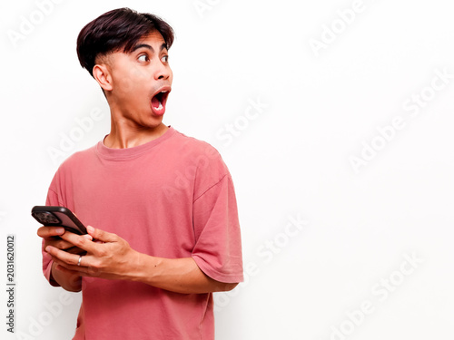 Фотография funny Excited young Asian man holding a smartphone and looking sideways in surprise, shocked by a commercial promotion or notification against a white studio background with copyspace