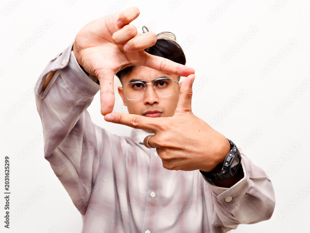 Naklejka premium Serious young Asian creative director making a finger frame gesture around his eyes, focusing closely at the camera to search for vision against a white studio background.