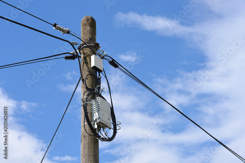 Vertical view of an old weathered wooden utility pole with a fiber optic distribution box and coiled black cables under a blue sky with white clouds