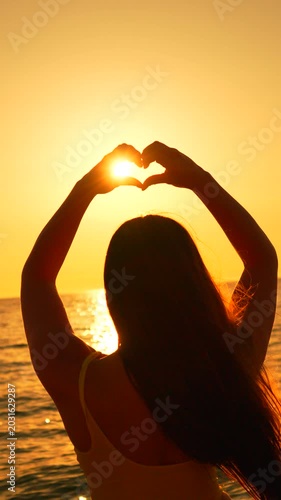 Silhouette heart sunset young woman makes a heart shape with her hands around the sun over ocean waves at the beach