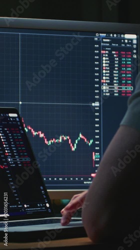 Unrecognizable man stock trader in home office. Back of male analyzes terminal, trades stocks on crypto market. Monitor and laptop display vivid candlestick or trading charts, price action. Evening