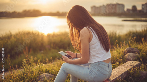 Slika na platnu Young woman sitting on a wooden bench by a tranquil lake at golden hour, engrossed in her smartphone