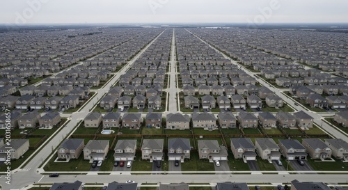 Aerial view of a vast, uniform suburban housing development with many identical houses arranged in a grid pattern under an overcast sky.