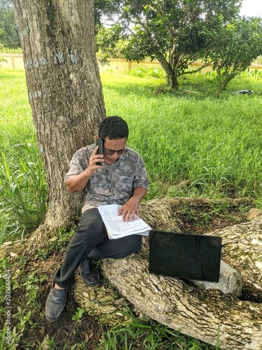 Man sitting under tree with laptop and phone.