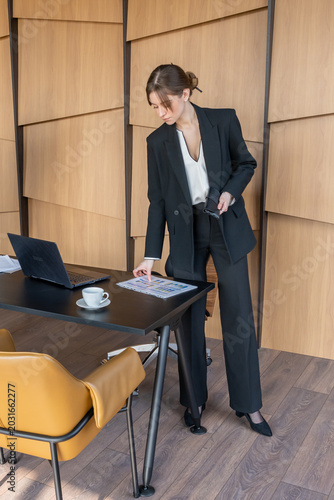Photos Businesswoman in a stylish pantsuit standing next to a modern office desk, revie