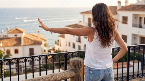 Tableau sur toile Rear view photograph of a female traveler with outstretched arm admiring the Mediterranean coast and charming town