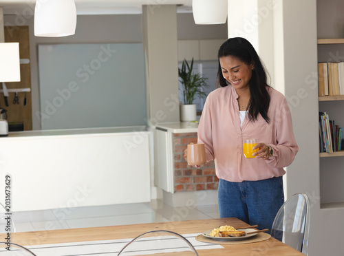 Asian woman standing at dining table holding ceramic mug and orange juice, plate nearby, copy space