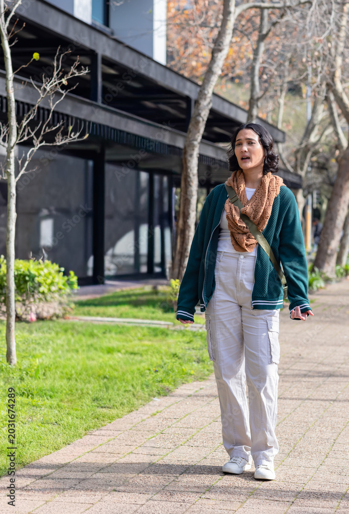 Fototapeta premium Adult woman walking along paved path beside building, holding phone and wearing green crossbody bag