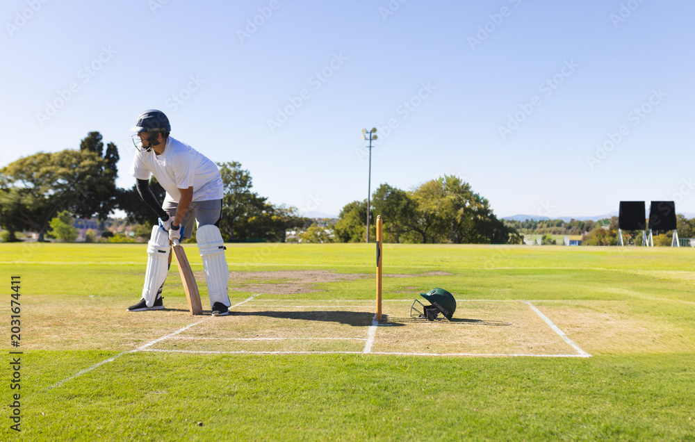 Obraz premium Male batter standing at crease by stumps on cricket ground holding wooden bat near spare helmet