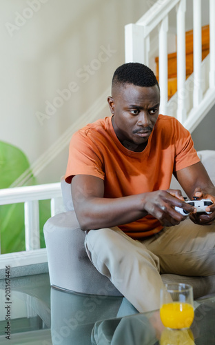 African American man leaning forward holding controller at home wearing orange tee with glass of OJ