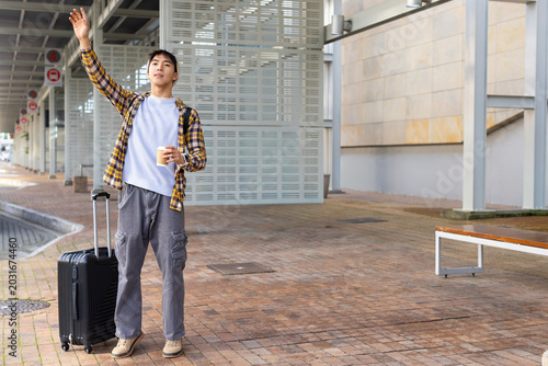 Adult Asian man raising arm while holding coffee cup beside black suitcase under canopy, copy space