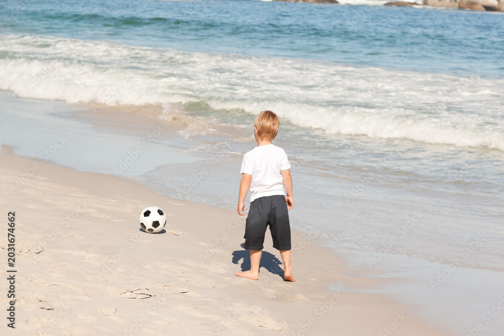 Obraz premium Boy child standing watching ocean at beach in white tee and dark shorts with soccer ball