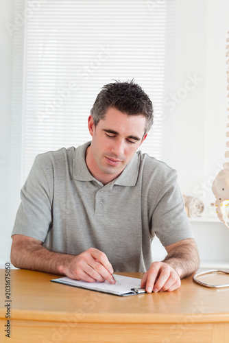 Принти на полотні Man writing on clipboard at desk in bright room with closed blinds, wearing gray