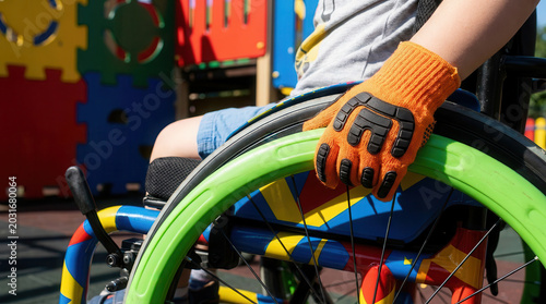 Child's hands gripping a colorful wheelchair wheel while seated, vibrant playground equipment visible in the background, showcasing an accessible play environment
