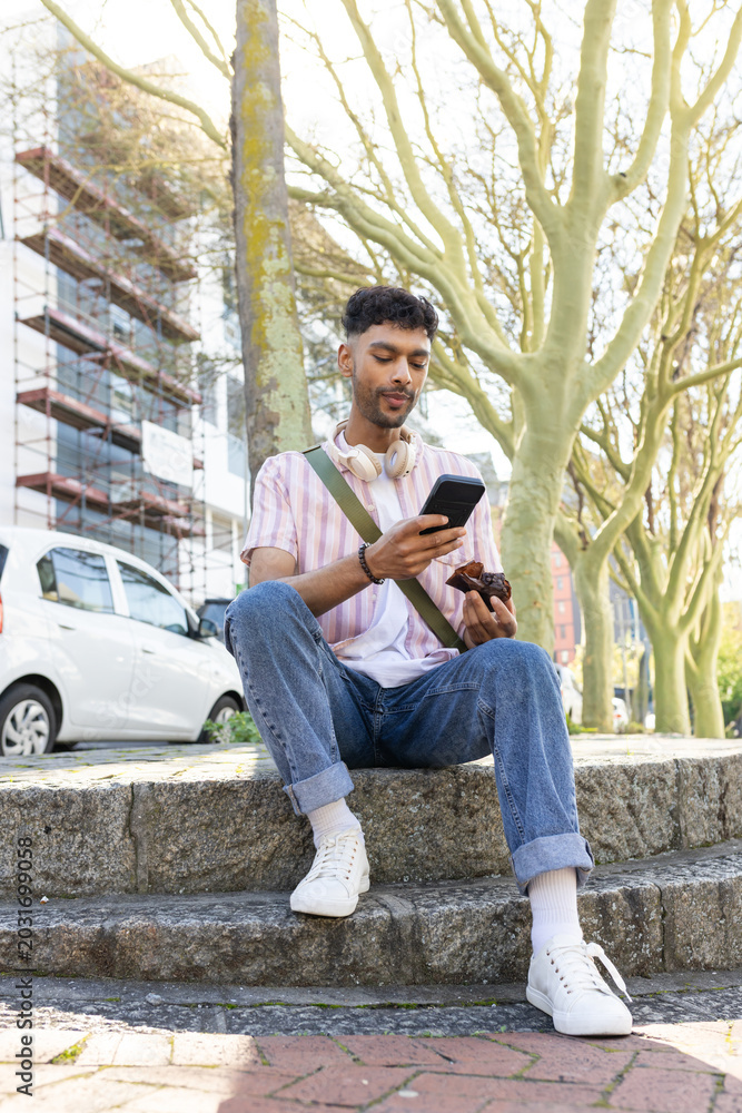 Obraz premium Man sitting on bench scrolling smartphone and eating pastry with headphones at tree-lined street