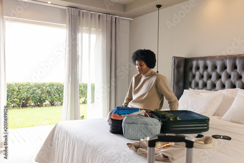 Mid adult African American female packing open suitcase on bed in beige sweater, pants, copy space
