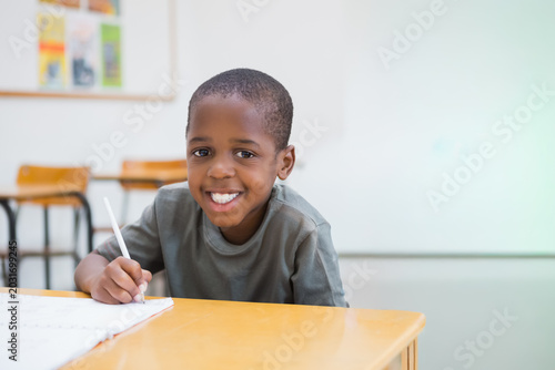 African American child sitting in classroom at wooden desk writing with pencil smiling at camera
