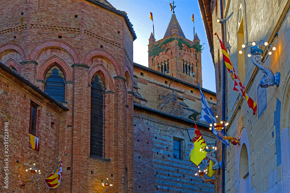 Naklejka premium SIENA, ITALY, April 24, 2026 : San Clemente in Santa Maria dei Servi and a small street decorated with flags