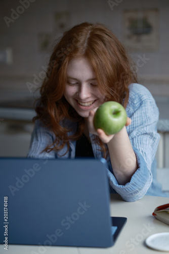 computer on a table. A teenage girl with beautiful red hair happily works on a computer, holding a green apple in her hand. She looks at the screen and communicates online. A new modern blue computer 