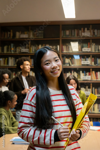 Young female student holding a folder and pen in a library with her classmates