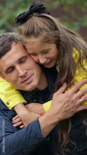 White Father And Daughter Hugging Outdoors, Smiling Girl With Long Brown Hair And Black Bow Perched On Fathers Back In Yellow Jacket, Gentle Embrace, Candid Emotional Moment In Leafy Park Conveying