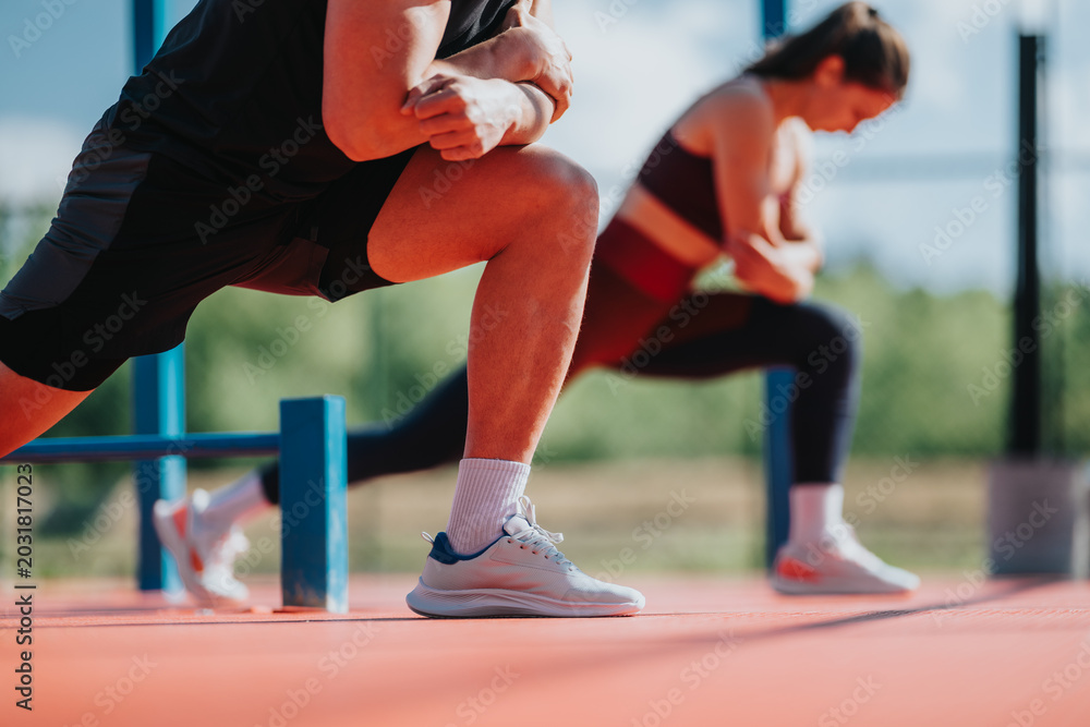 Fototapeta premium Man and woman perform forward lunges while stretching and exercising outdoors. A close-up scene of partners doing coordinated fitness training on a court.