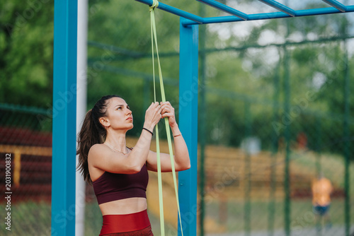 Photography A young woman secures a resistance band to outdoor monkey bars while focusing on her exercise setup