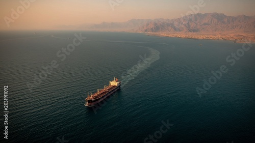 A gorgeous low-light photograph of a cargo ship passing through the congested Strait of Hormuz, with the Iranian shoreline in the distance