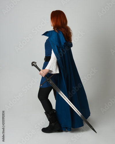 full length portrait of female red head model wearing medieval fantasy warrior historical costume with blue cloak. silhouette back view figure walking away and holding sword weapon, studio background
