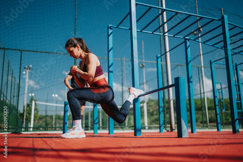 Fotografie A fit woman does a Bulgarian split squat with her rear foot elevated on a bar at an outdoor calisthenics park